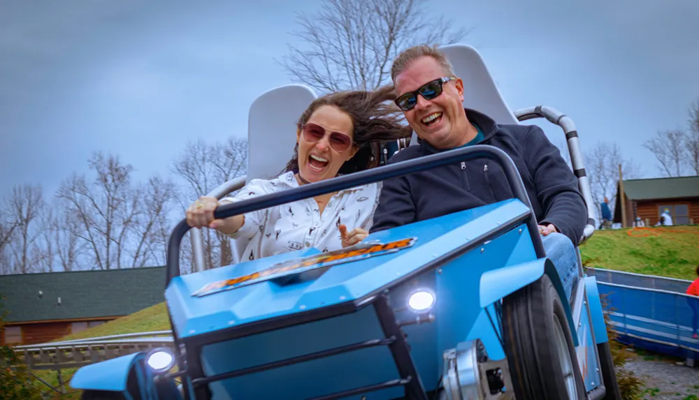 Two people are joyfully experiencing a ride in a blue roller coaster car with dynamic expressions and windswept hair