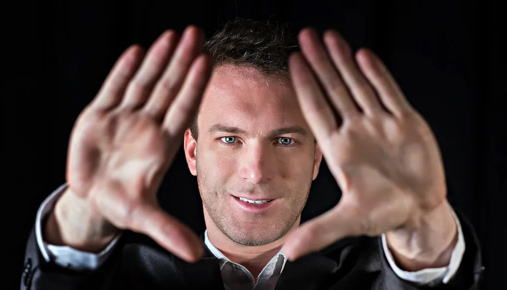 A man in a black suit is framing his face with his hands against a black background