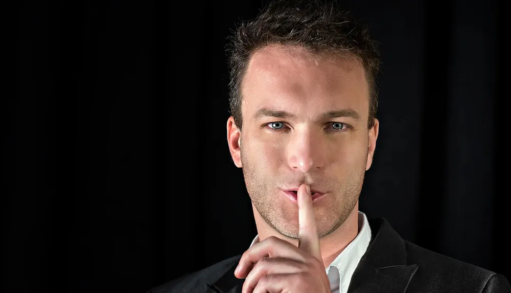 A man in a black suit jackets is holding his index finger to his lips in a gesture commonly used to indicate silence or secrecy