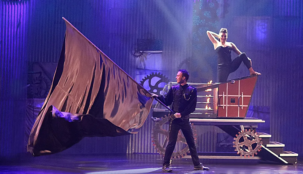 A performer waves a large flag while another performer strikes a pose atop a moving mechanical apparatus on stage set against a backdrop with cogs adding an industrial or steampunk vibe to the scene