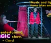 A dynamic scene from a magic show features a woman in a red dress interacting with a performer chained to a frame set against a festive backdrop with quotes praising the entertainment