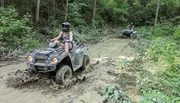 Two people are riding all-terrain vehicles through a muddy forest trail.