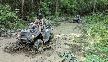 Two people are riding all-terrain vehicles through a muddy forest trail.