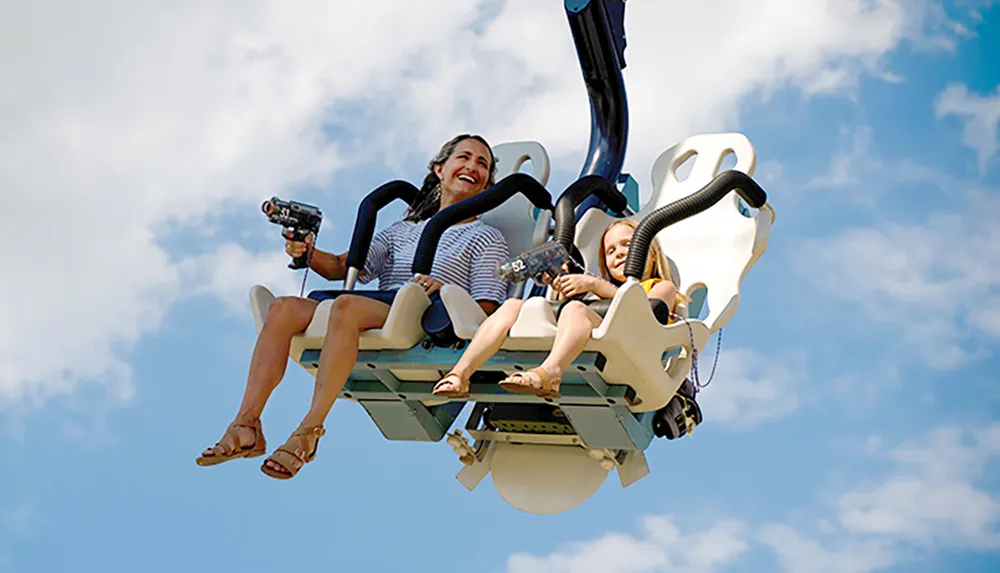 Two individuals likely a mother and child appear thrilled while riding on a suspended amusement park ride against a backdrop of a clear blue sky