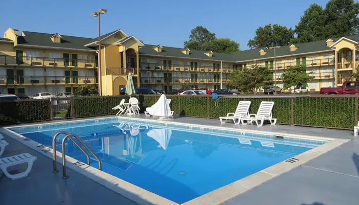 The image shows a clear outdoor swimming pool with lounge chairs in front of a multi-story motel complex on a sunny day