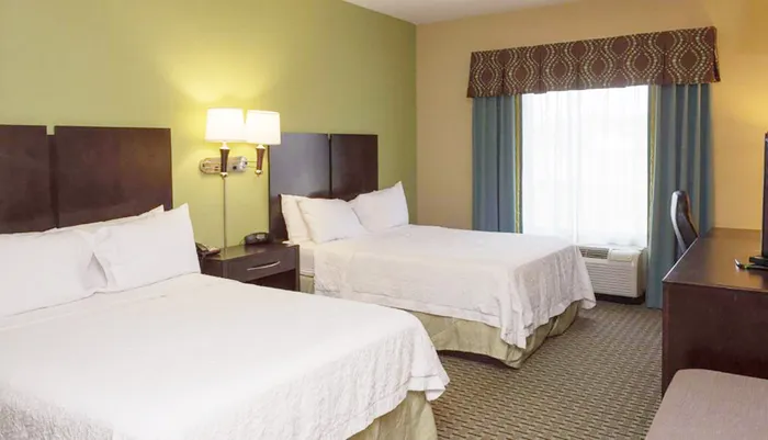 The image shows a tidy hotel room with two double beds a nightstand with a lamp a window with curtains and a wall-mounted television