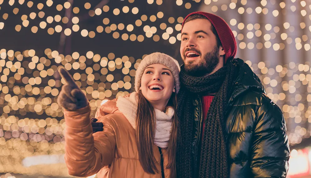 A joyful couple is enjoying a festive night out surrounded by sparkly lights while the woman points at something interesting outside the frame