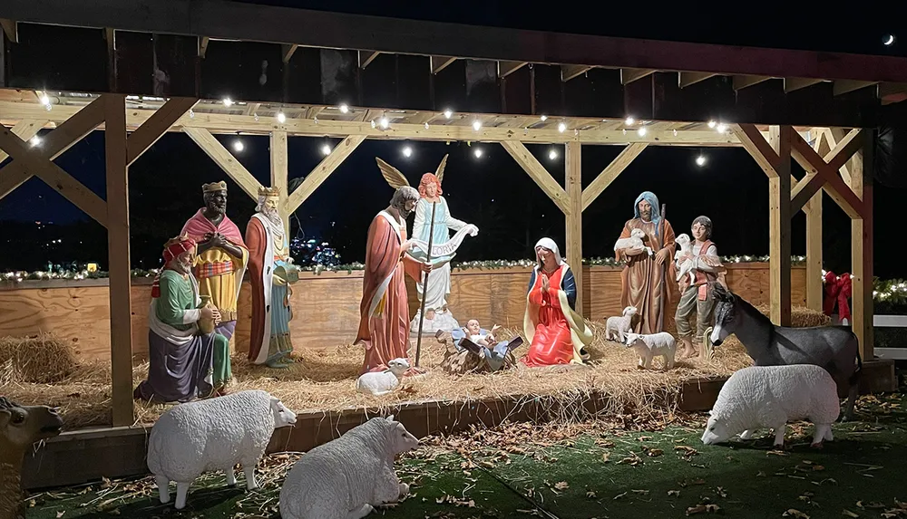 This image depicts a nativity scene with life-size figures including Mary Joseph the baby Jesus the three wise men shepherds an angel and various animals set within a wooden stable structure against a night sky backdrop