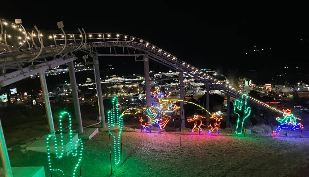 The image shows a nighttime view of a festive outdoor setting with an illuminated alpine coaster track in the foreground and colorful light displays shaped like a cactus and reindeer with city lights sprawling in the background