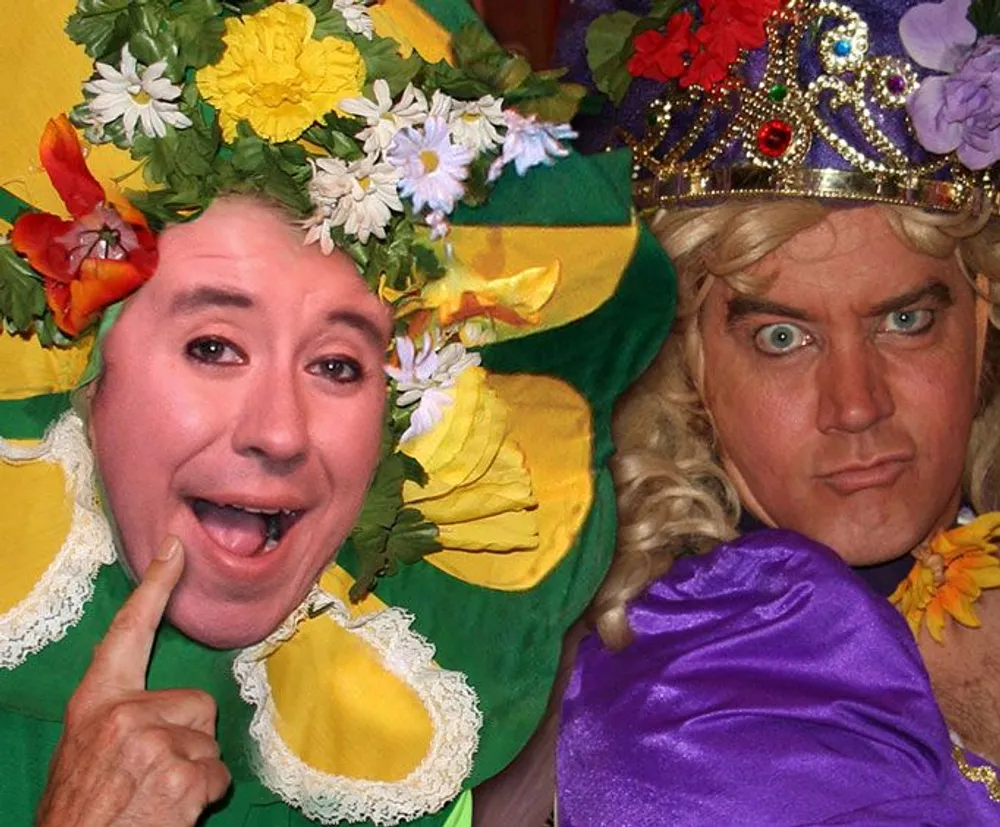 Two people wear humorous costumes with exaggerated floral hats and expressive makeup one smiling and pointing at their cheek and the other with a mock stern look
