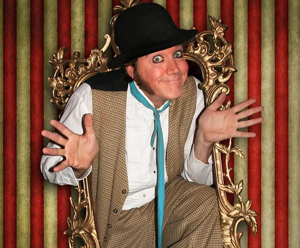 A person dressed in a bowler hat and vintage attire is striking a playful pose through a golden ornate picture frame against a red and gold striped backdrop