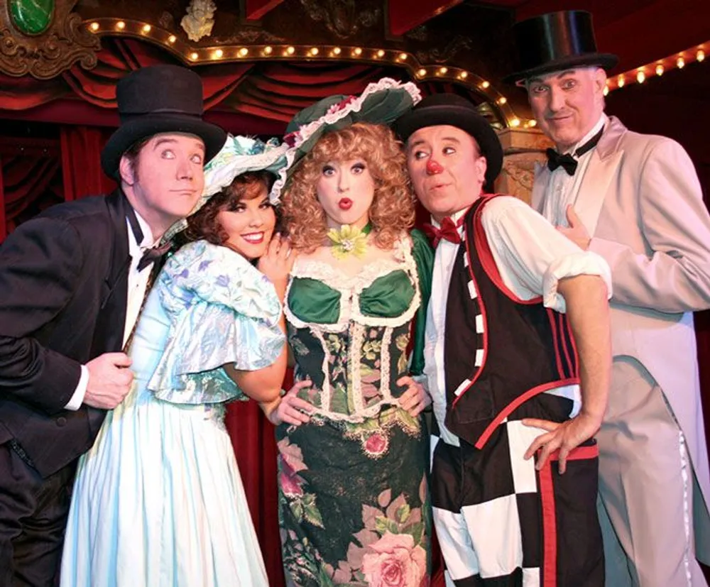 Five actors in elaborate historical costumes pose playfully against a theatrical backdrop suggesting a Vaudeville or old-time stage performance