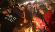 A group of people are gathered around a lantern outdoors at night, paying attention to a person who appears to be leading a tour or telling a story.