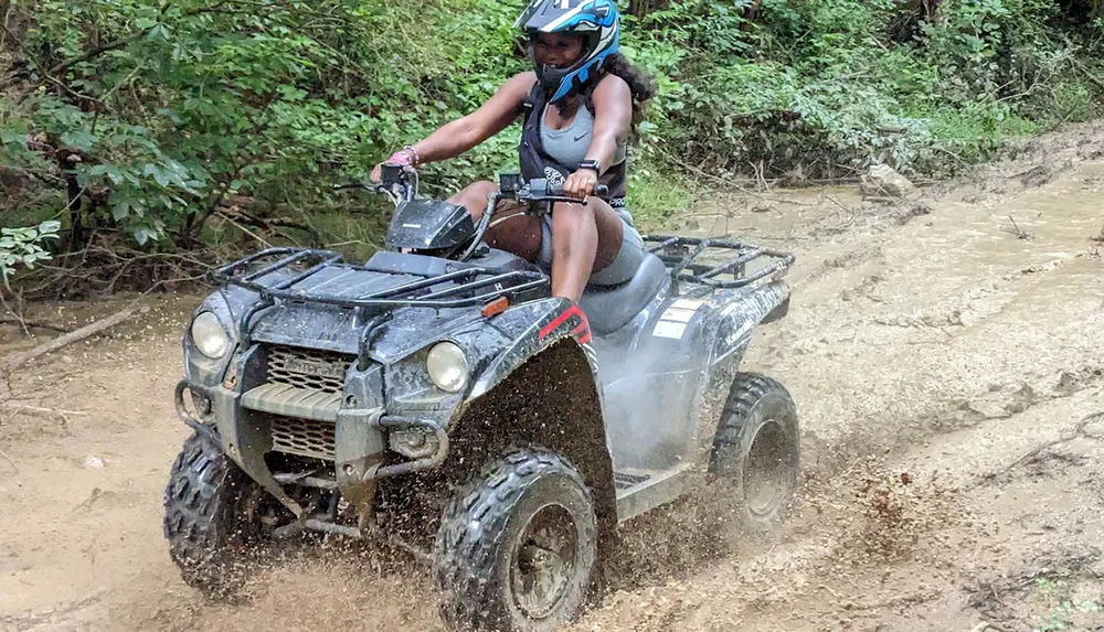 A person wearing a helmet is actively riding an ATV through a muddy trail splashing dirt around