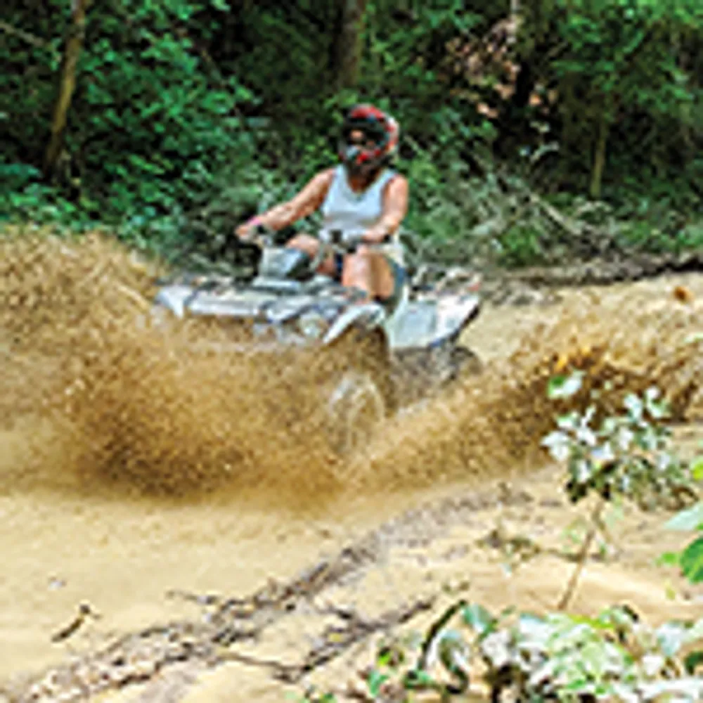 A person wearing a helmet is riding an ATV through a muddy trail in a forested area