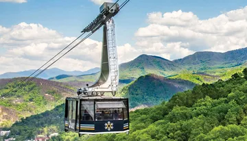 A cable car with a logo on it traverses a scenic landscape with lush green mountains and patches of residential areas below.