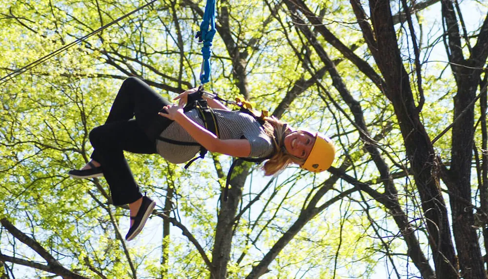 A person is wearing a helmet and harness while ziplining amongst trees on a sunny day