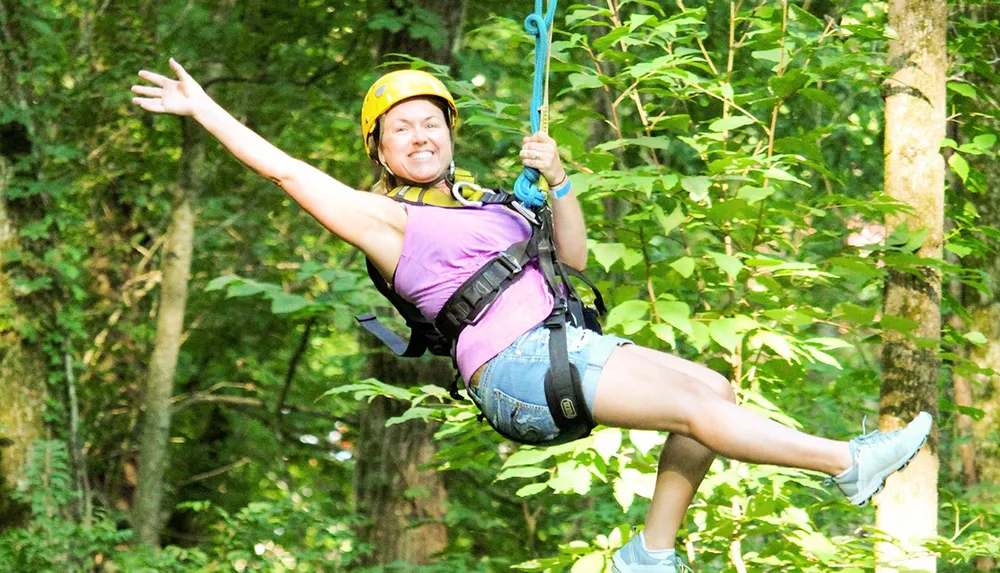 A person is joyfully zip-lining through a forest wearing a helmet and a harness for safety