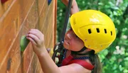 A young person wearing a yellow helmet is focused while climbing a wooden wall with green handholds.