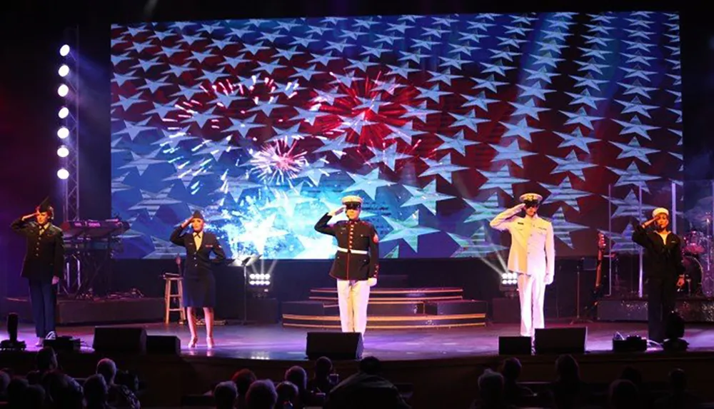 Military personnel in uniform are saluting on stage in front of a large American flag backdrop