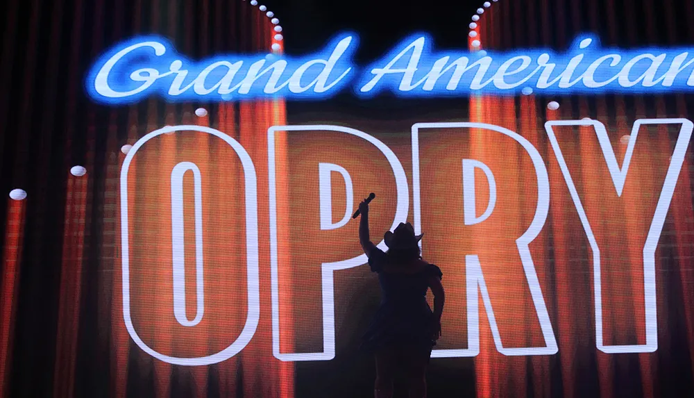 A silhouette of a performer holding a microphone stands in front of a large illuminated sign that reads Grand American Opry