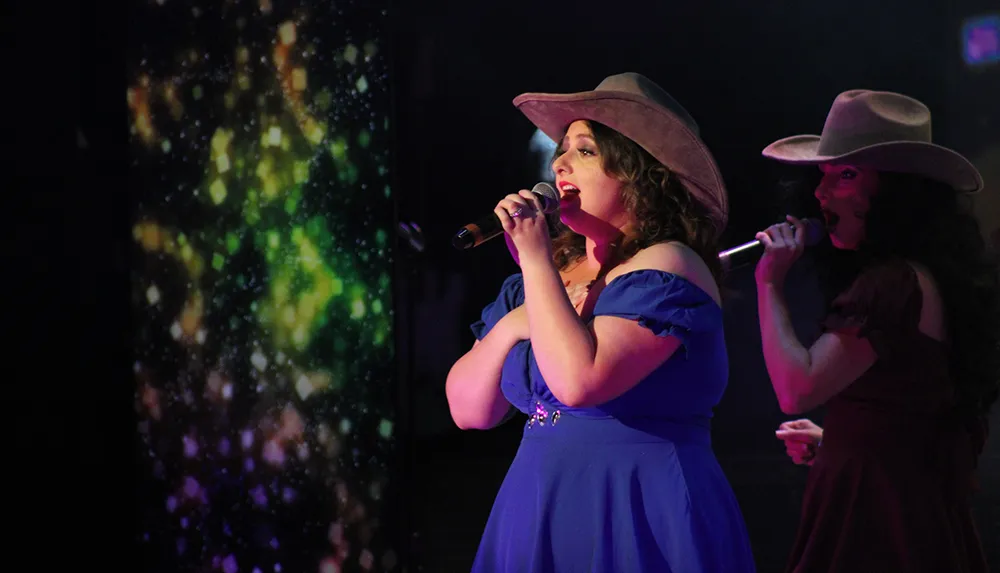 Two women wearing cowboy hats are performing on stage one in a blue dress and the other in maroon against a backdrop of colorful lights