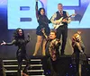 A group of performers is posing energetically on stage in front of a large sign with a positive quote about their singing and an inset image of a meal featuring chicken mashed potatoes and green beans