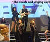 A group of performers is posing energetically on stage in front of a large sign with a positive quote about their singing and an inset image of a meal featuring chicken mashed potatoes and green beans