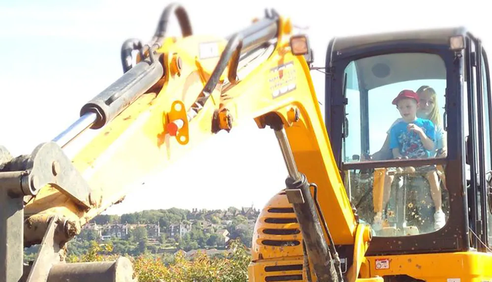 A child and an adult are sitting inside the cab of an excavator with the child appearing curious or excited
