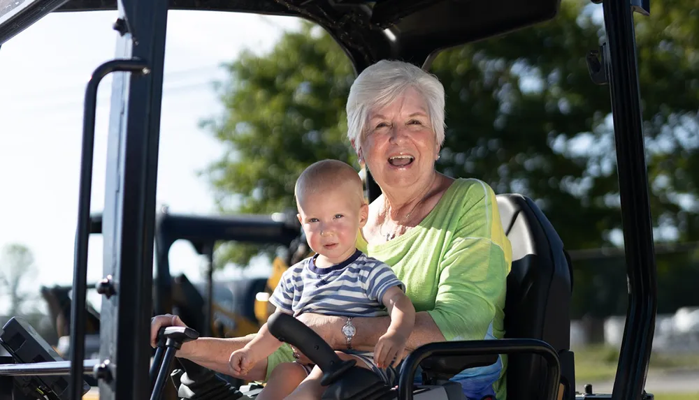 An elderly woman and a young child are smiling while sitting together on a piece of outdoor equipment perhaps a tractor