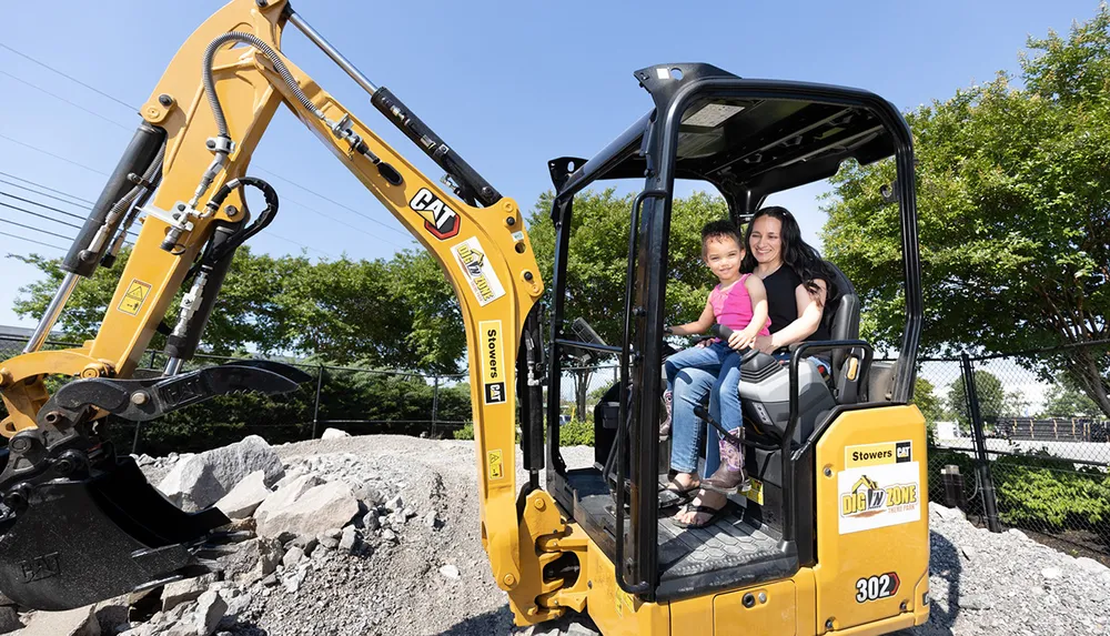 A woman and a child are sitting inside the cabin of a yellow CAT mini excavator smiling