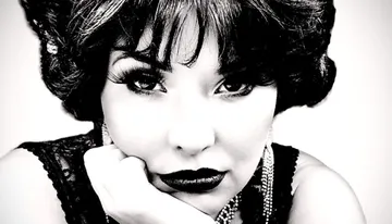 The image features a close-up, black-and-white portrait of a woman with a vintage hairstyle, dark lipstick, and intense gaze, resting her chin on her hand.