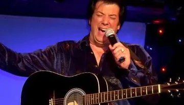A person is passionately singing into a microphone with a guitar in the foreground and colorful lights in the background.