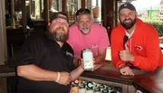 Three men are smiling at the camera, holding a jar, inside a rustic setting.