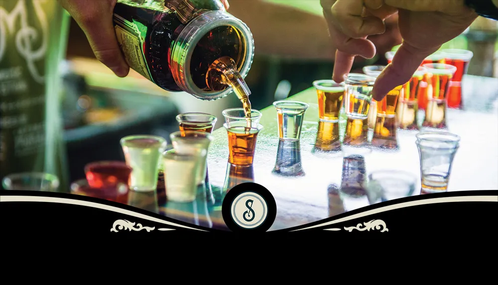 A person is pouring a dark liquid from a bottle into an array of shot glasses on a reflective surface with another hand pointing at one of the glasses