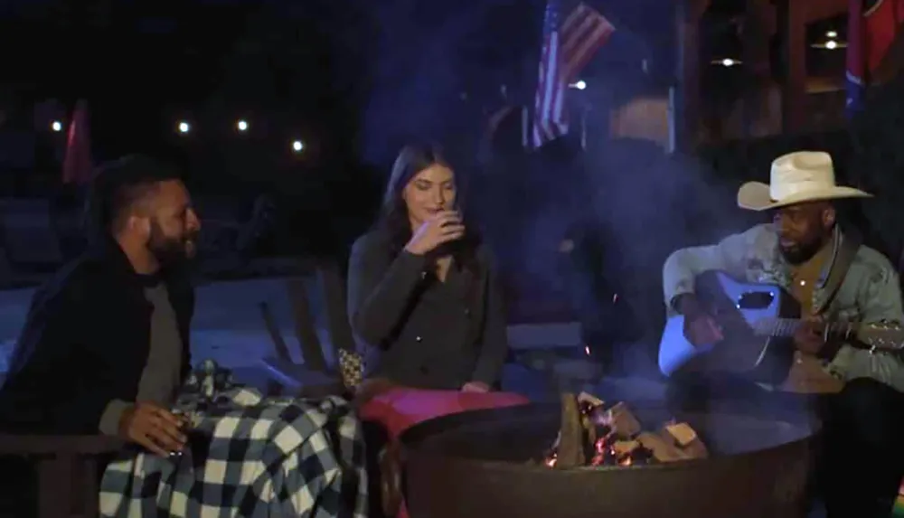 Three people are sitting around a fire pit at night with one person playing a guitar and the other two drinking
