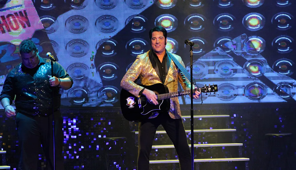 A performer wearing a gold blazer plays a guitar on stage accompanied by another person in a glittery outfit
