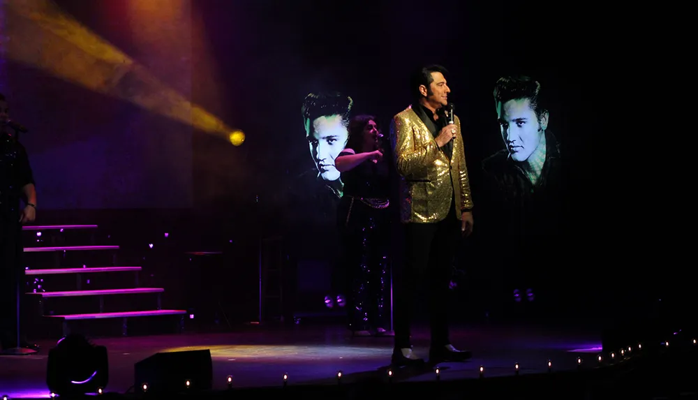 A male performer in a gold jacket sings on stage with two large projected images of a famous musician in the background
