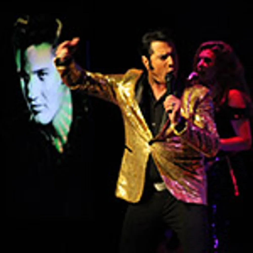 A performer in a gold jacket dances energetically on stage with singers in the background and a large dog illustration on the screen behind them