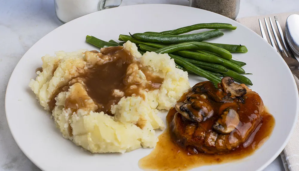 A plate of mashed potatoes with gravy green beans and a piece of meat topped with mushrooms and sauce