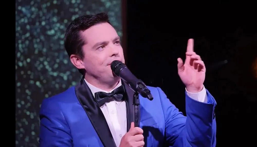 A man in a blue suit and bow tie is speaking or singing into a microphone gesturing with his hand