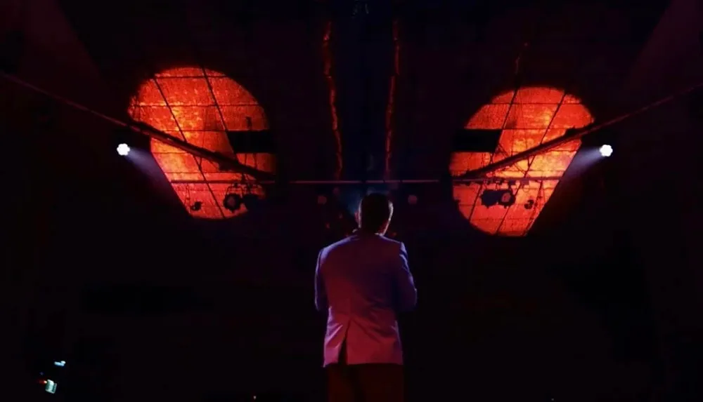 A person in a suit stands on stage facing an audience with large orange circles projected overhead