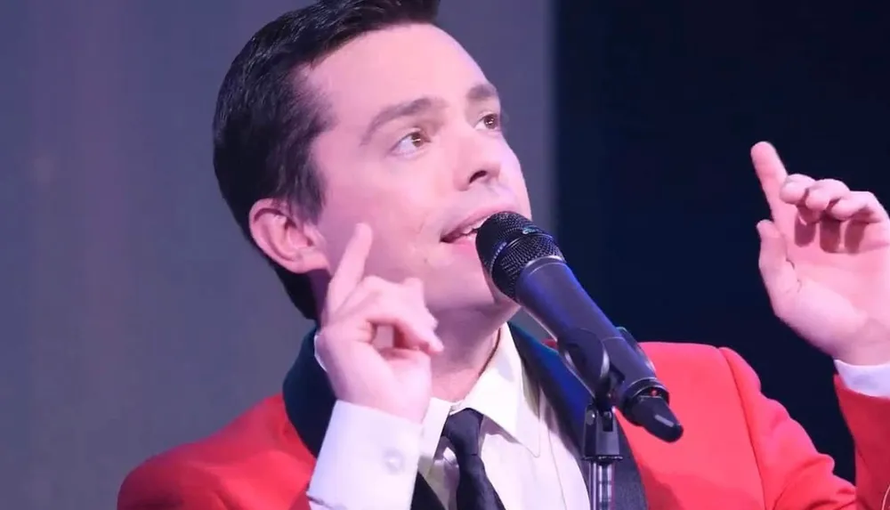 A person wearing a red jacket is singing into a microphone with their hands raised