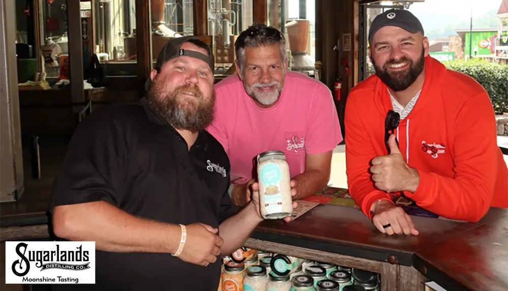 Three men are smiling at a moonshine tasting event at Sugarlands Distilling Co with one of them holding a jar of moonshine