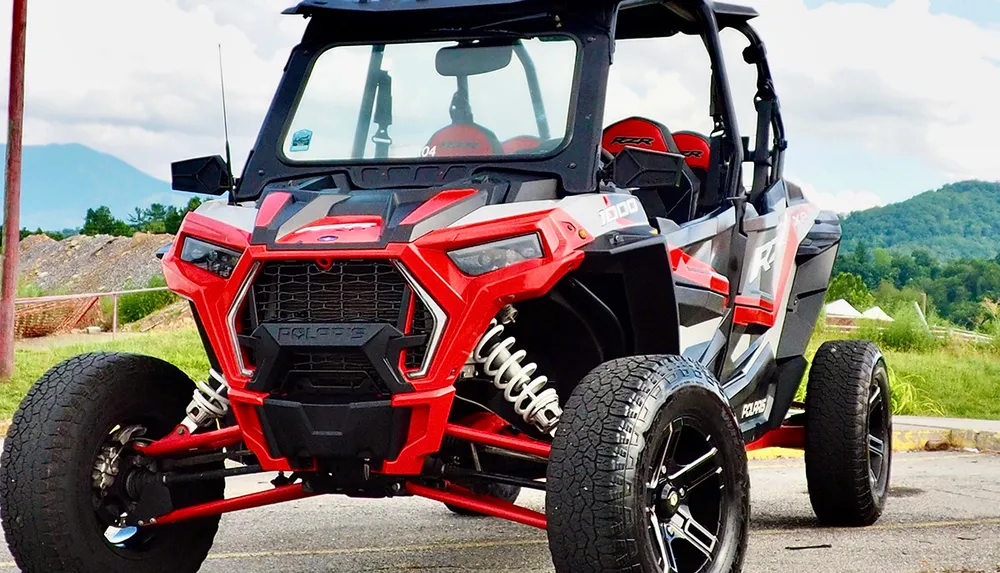 A red and black off-road vehicle is parked outdoors against a backdrop of green hills and a cloudy sky