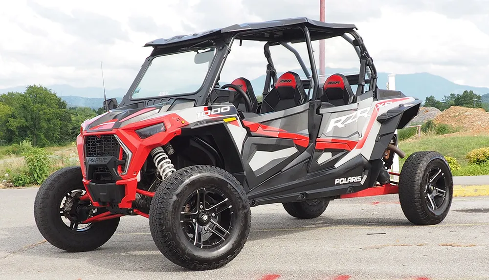 The image shows a red and black off-road vehicle likely a Polaris RZR parked on a paved area with greenery and mountains in the background