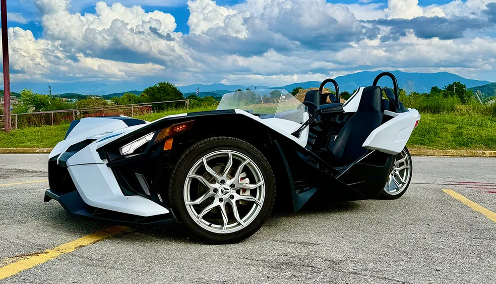 A white three-wheeled vehicle is parked on a road with a scenic mountain landscape in the background