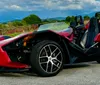 A red Polaris Slingshot three-wheeled vehicle is parked outdoors with a scenic backdrop of hills and cloudy skies