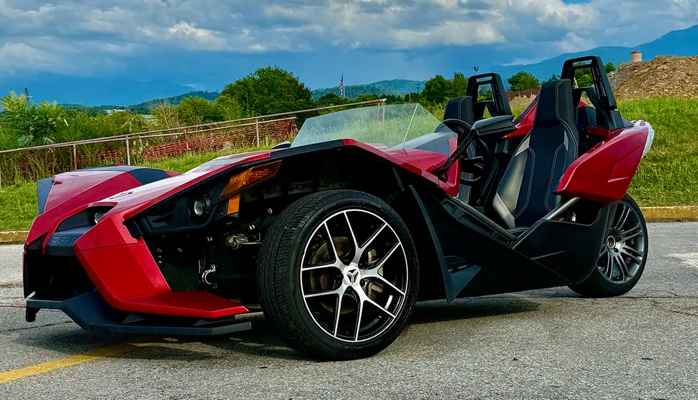 A red Polaris Slingshot three-wheeled vehicle is parked outdoors with a scenic backdrop of hills and cloudy skies