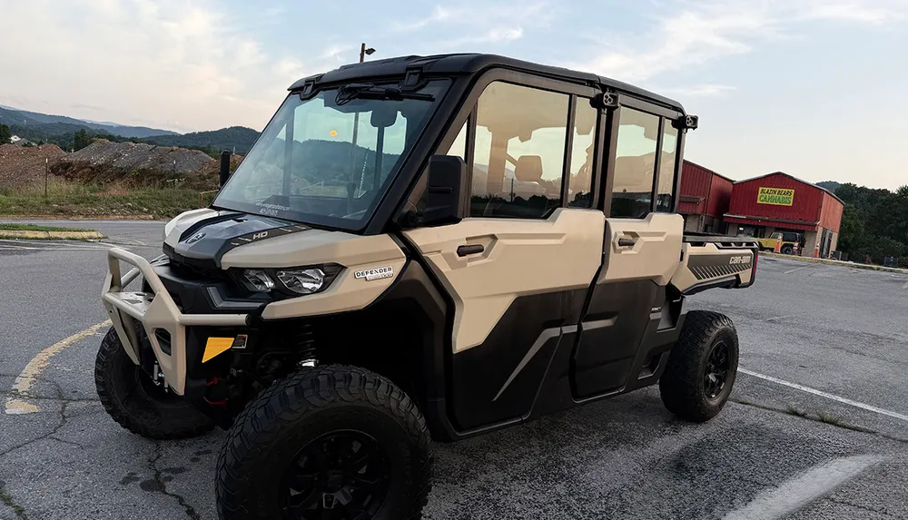 A tan and black Can-Am Defender side-by-side utility vehicle is parked in an outdoor setting near a red building